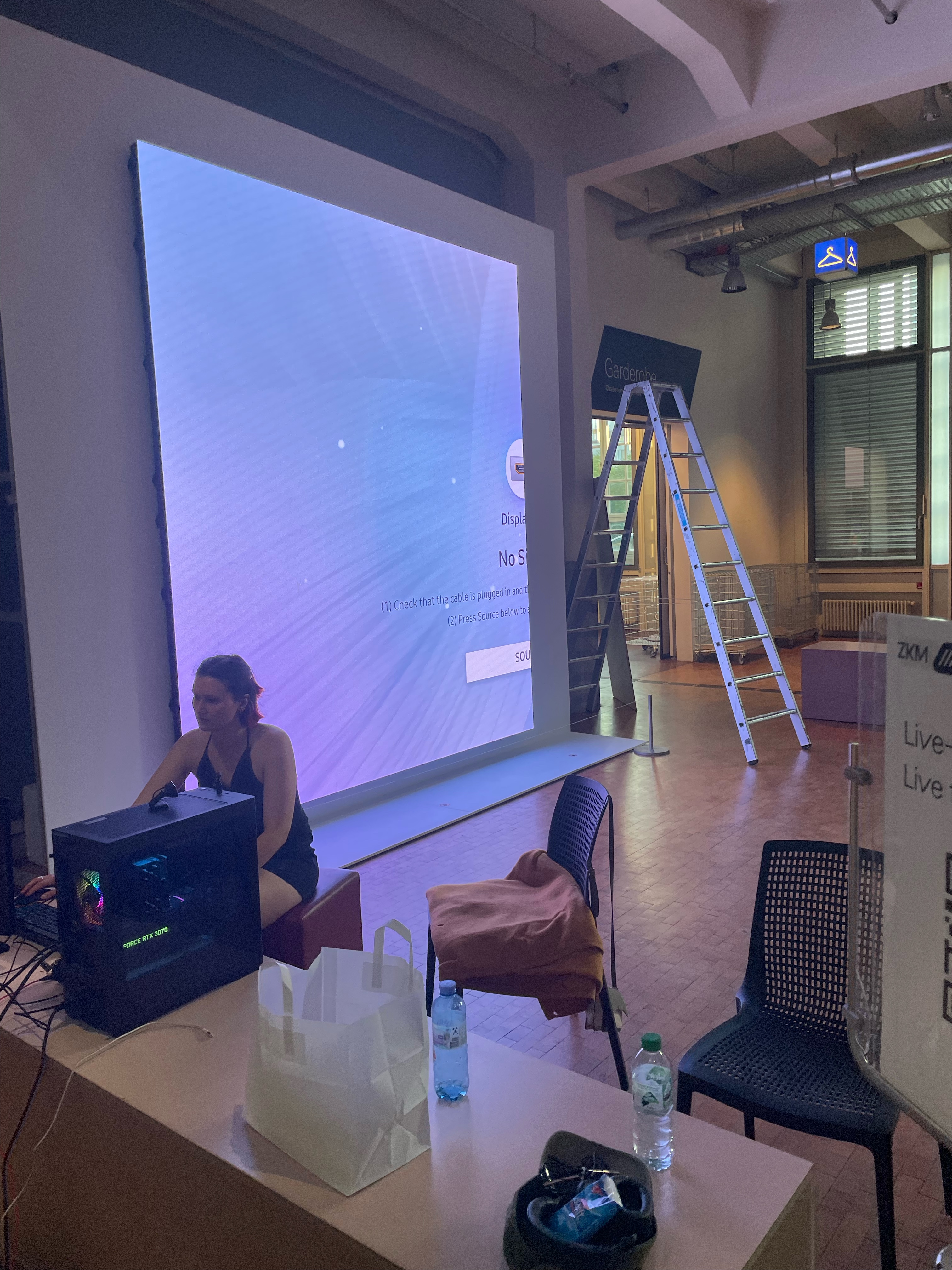 Katharina arbeitet an ihrem Laptop. Um sie herum liegt verteilt technisch. Im Hintergrund ist eine Leiter aufgebaut und die LED Wand zeigt ein Standbild.