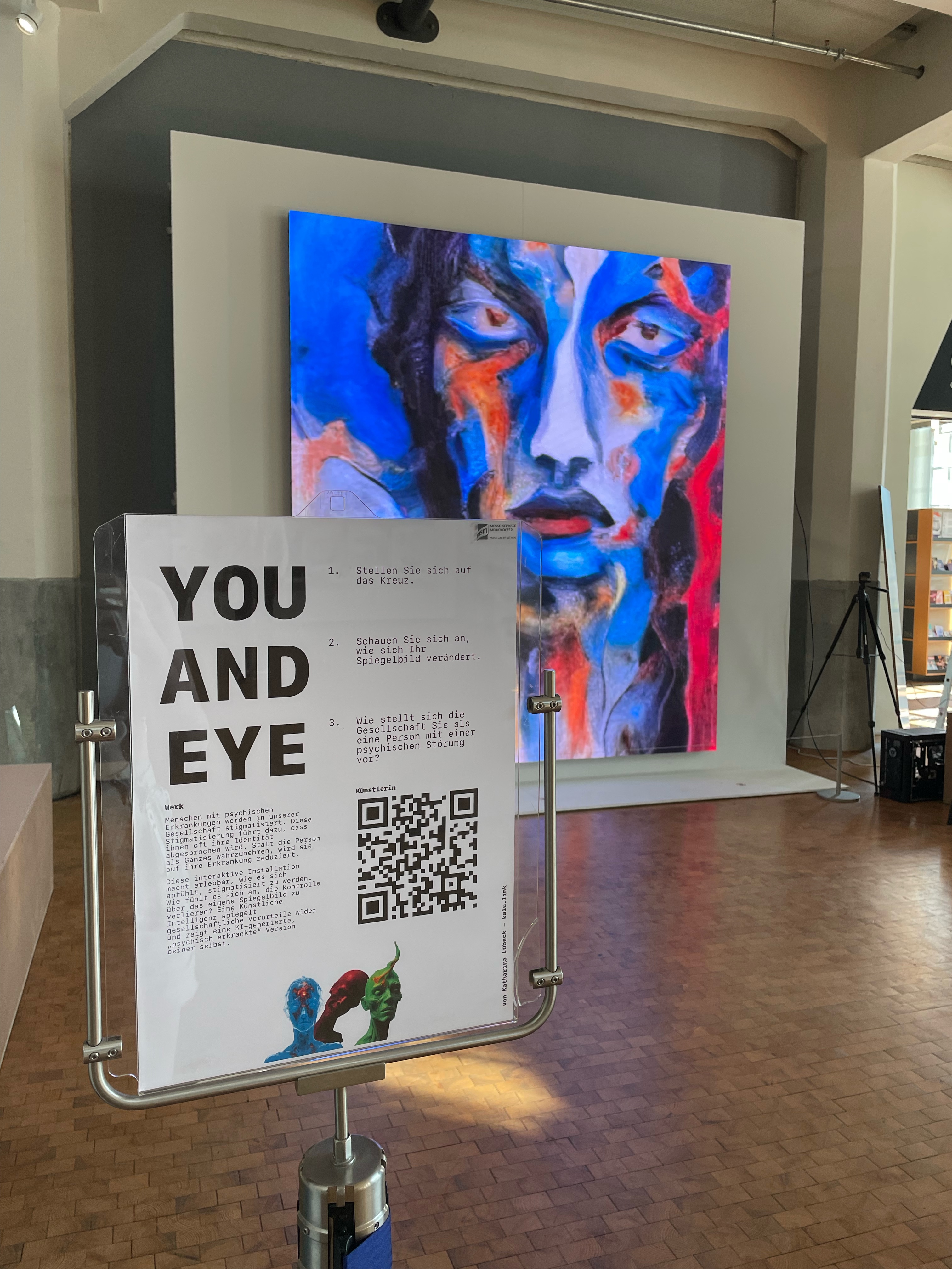 Die LED Wand zeigt ein buntes Gesicht, das aussieht wie gezeichnet. Davor steht ein Informationsschild was über die Ausstellung 'You and Eye' informiert.