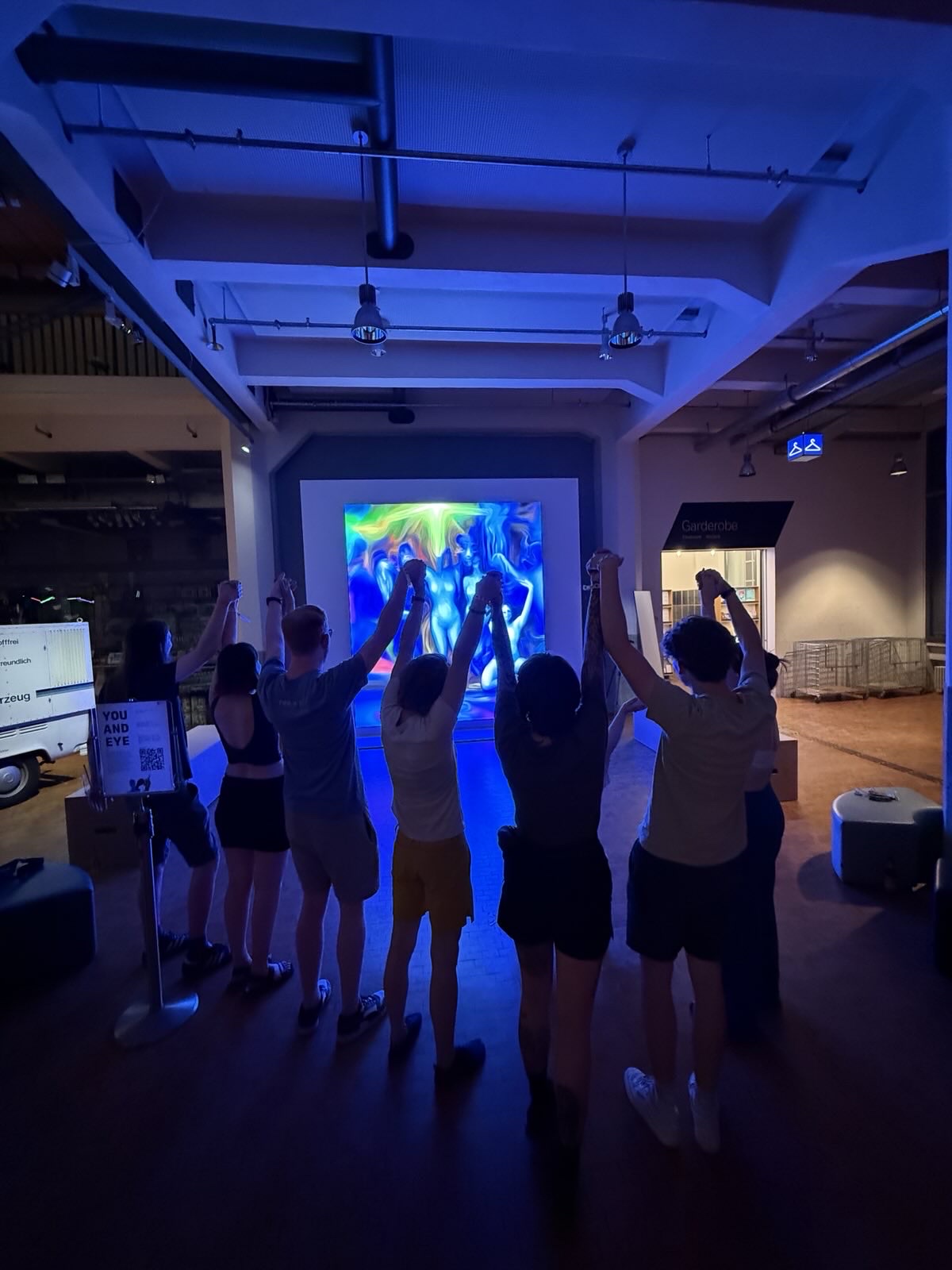 Eine Gruppe von sieben Menschen, hält sich an den Händen und streckt diese nach oben. Auf dem LED Bildschirm wird die Personengruppe verzerrt und bunt dargestellt.