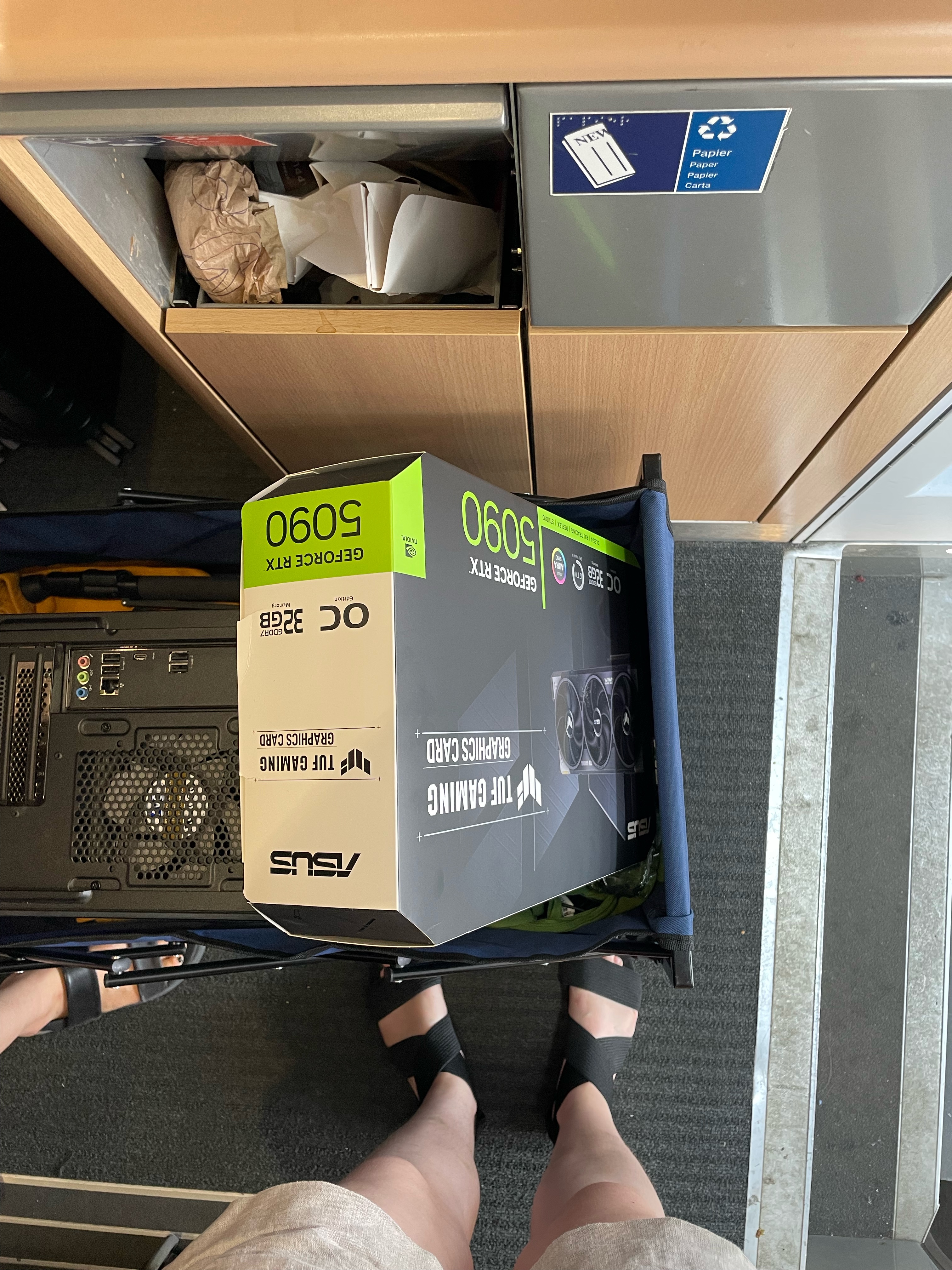 Vogelperspektive auf einen Handwagen, in dem sich ein kompletter Desktop-Rechner, eine Grafikkarte gtx 5090 und ein Stativ befindet. Der Hintergrund gibt zu erkennen, dass all das sich gerade im Eingangbereich eines ICEs befindet.