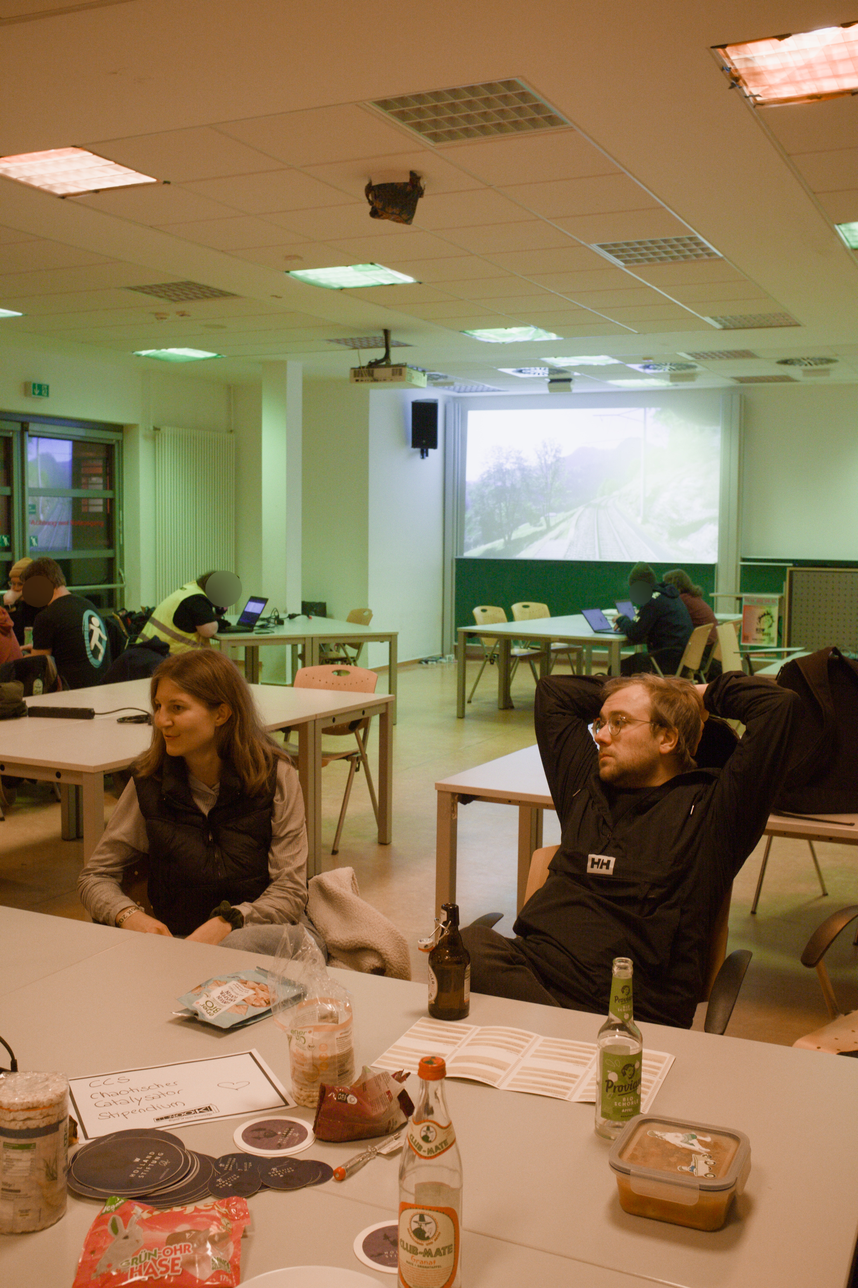 Zwei Persoen sitzen im bunt beleuchteten Kellerraum und unterhalten sich. Auf dem Tisch vor Ihnen liegt allerlei Krimskrams.