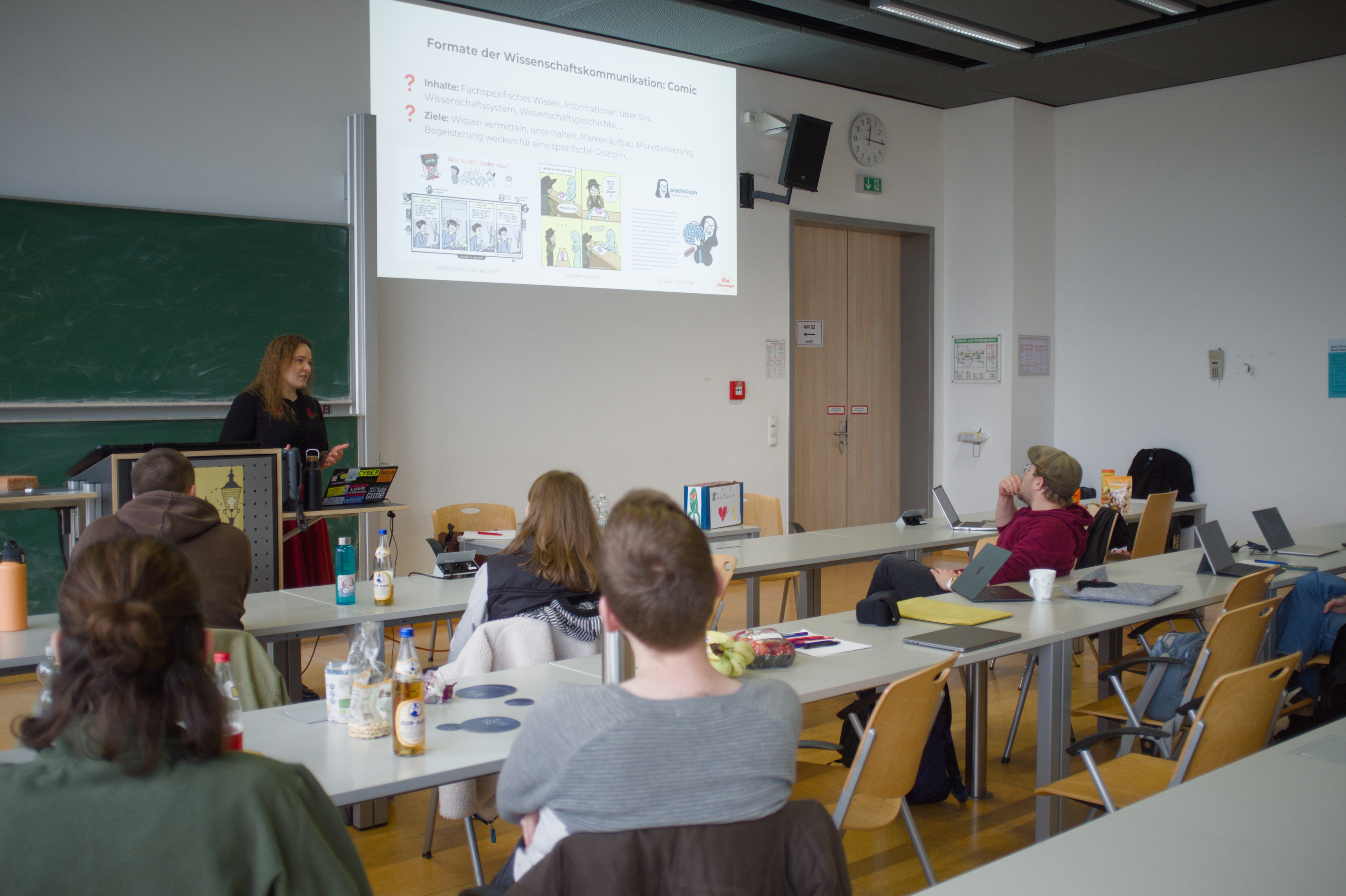 Lea hält einen Vortrag in einem Uniraum. Hinter ihr kann man die Vortragsfolien zum Thema Medien der Wissenschaftskommunikation erkennen. Vor ihr sitzen einige Personen und schauen den Vortrag an.