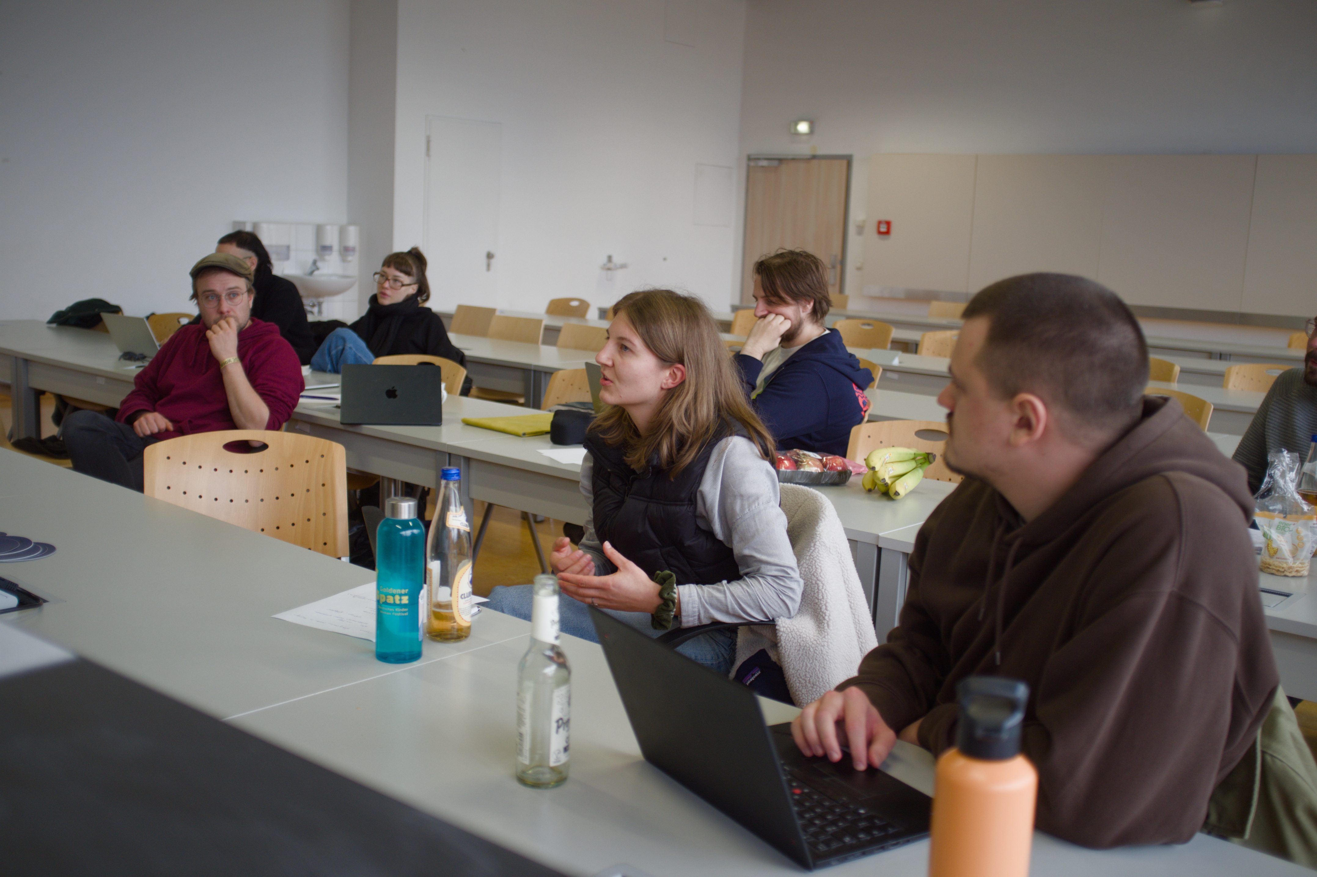 Hinter den Vorlesungstischen sitzen einzelne Personen und schauen an der Kamera vorbei. Eine junge Frau spricht gerade, die anderen höhren ihr zu.