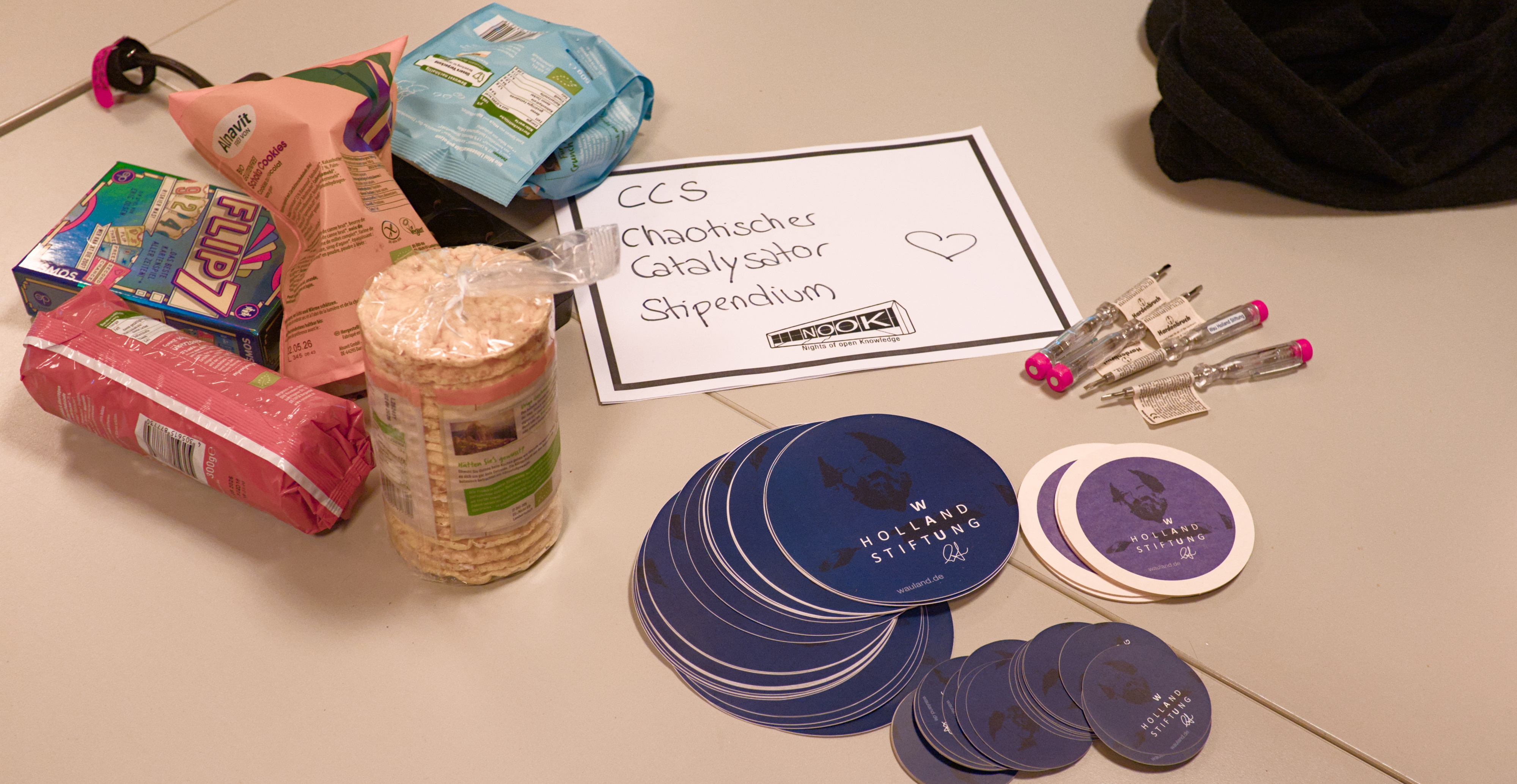 Auf einem Tisch liegt ein Zettel mit der Aufschrift 'CCS Chaotischer Catalysator Stipendium', daneben viele Snakcs und mehrere Stapel Wau Holland Stiftungs Sticker.