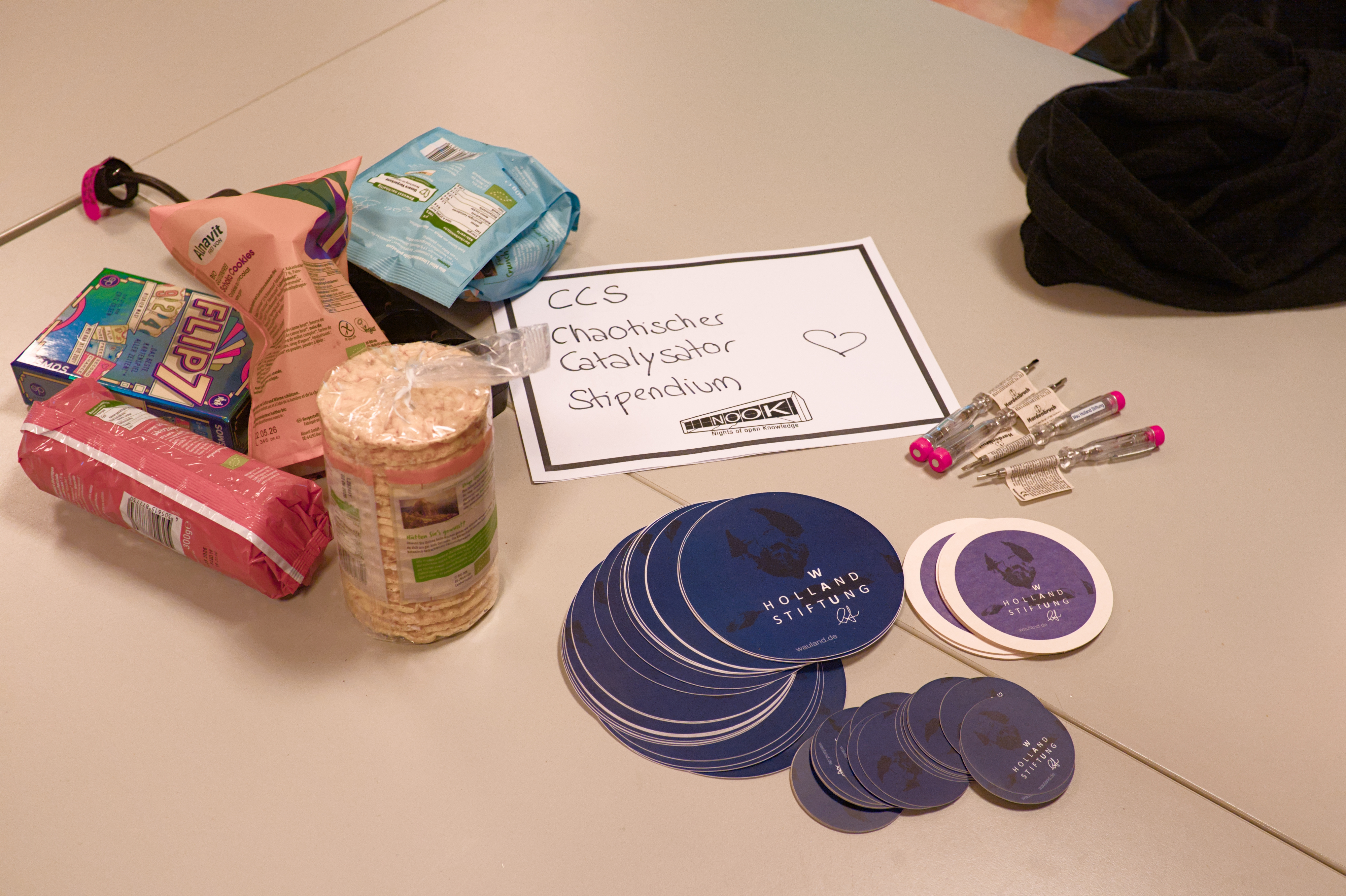 Auf einem Tisch liegt ein Zettel mit der Aufschrift 'CCS Chaotischer Catalysator Stipendium', daneben viele Snakcs und mehrere Stapel Wau Holland Stiftungs Sticker.