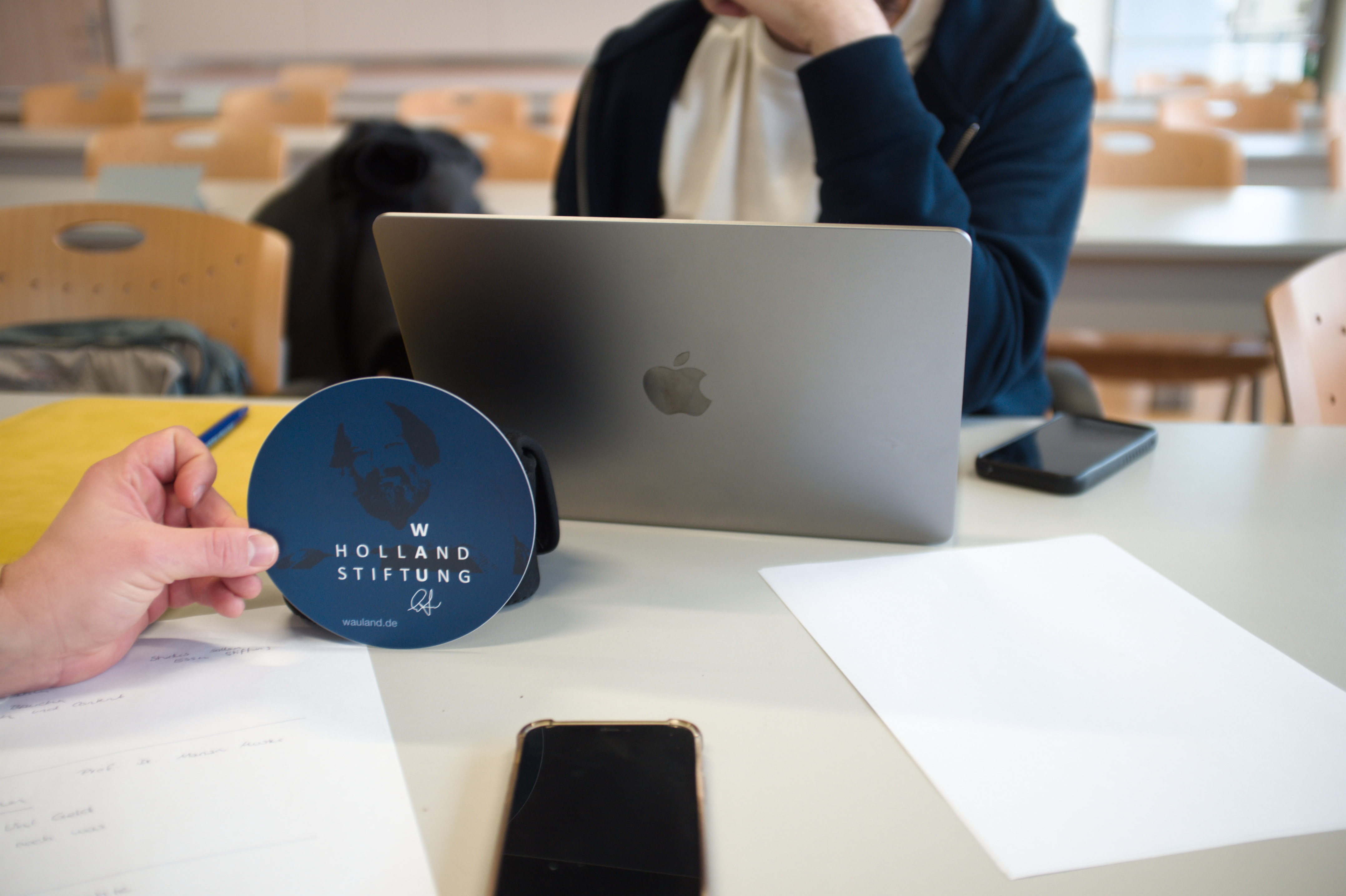 Eine Person die nicht klar erkennbar ist, sitzt hinter ihrem Laptop. Davor hält eine Hand einen Wau holland Sticker ins Bild.