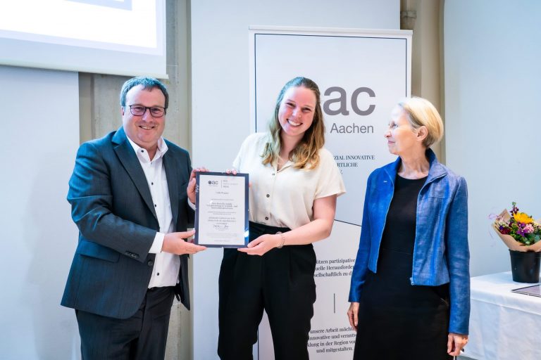 Laila hällt zusammen mit einem Mann im Anzug, stolz ihre Auszeichnung in der Hand. Beide lächeln in die Kamera. Rechts von ihnen steht eine Frau und schaut beide an.