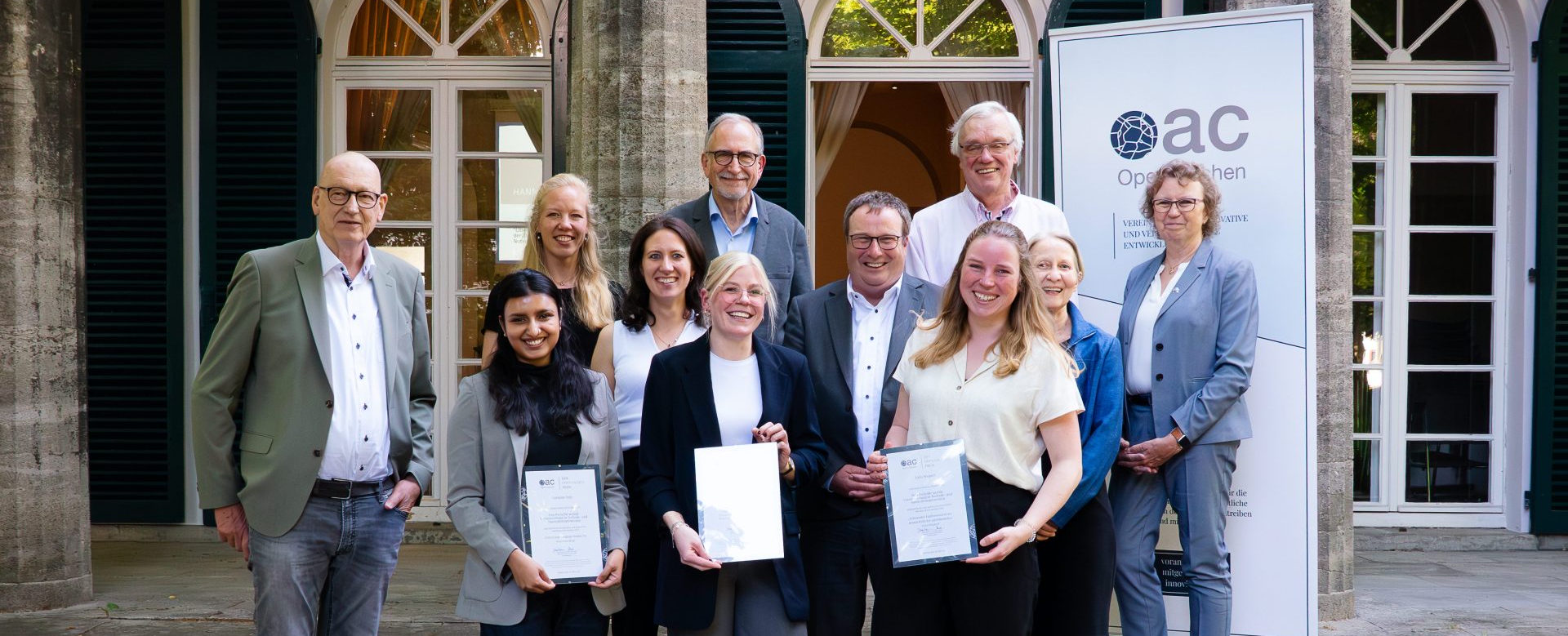 Drei ausgezeichnete Studentinnen posieren mit ihren Zertifikaten. Hinter ihnen stehen acht weitere Personen. Alle lächeln in die Kamera.