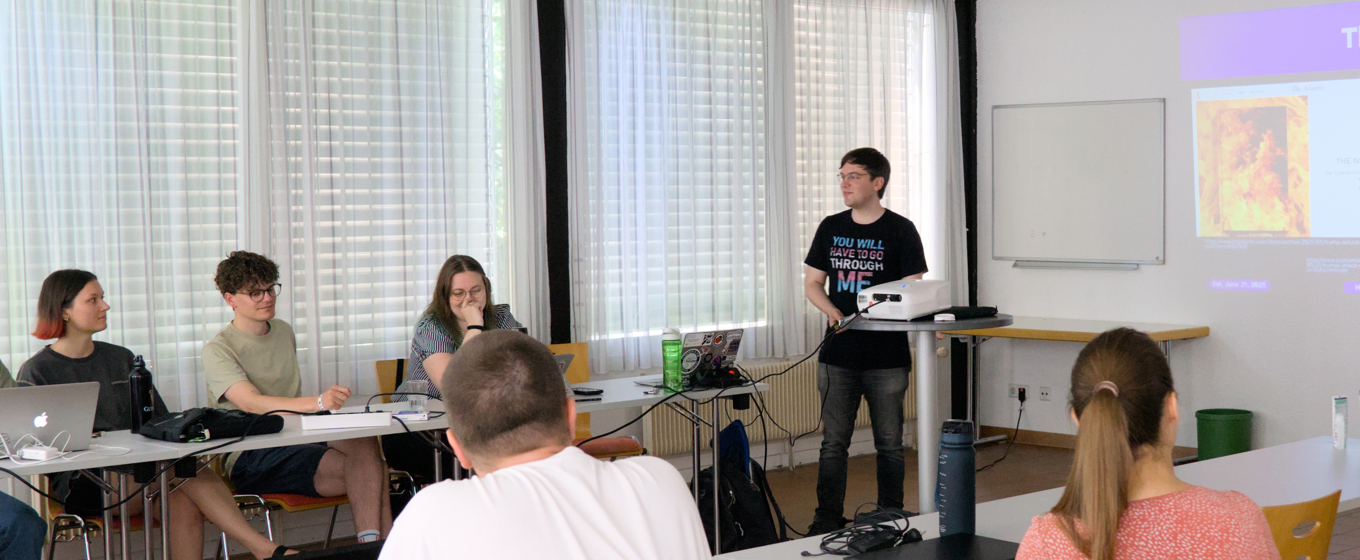 Henrik Schönemann steht neben einem Projektor und präsentiert nicht zu erkennende Folien. Daneben sitzen hinter Tischen mit aufgeklappten Laptops und Wasserflaschen, mehrere teilnehmende und schauen in seine Richtung.