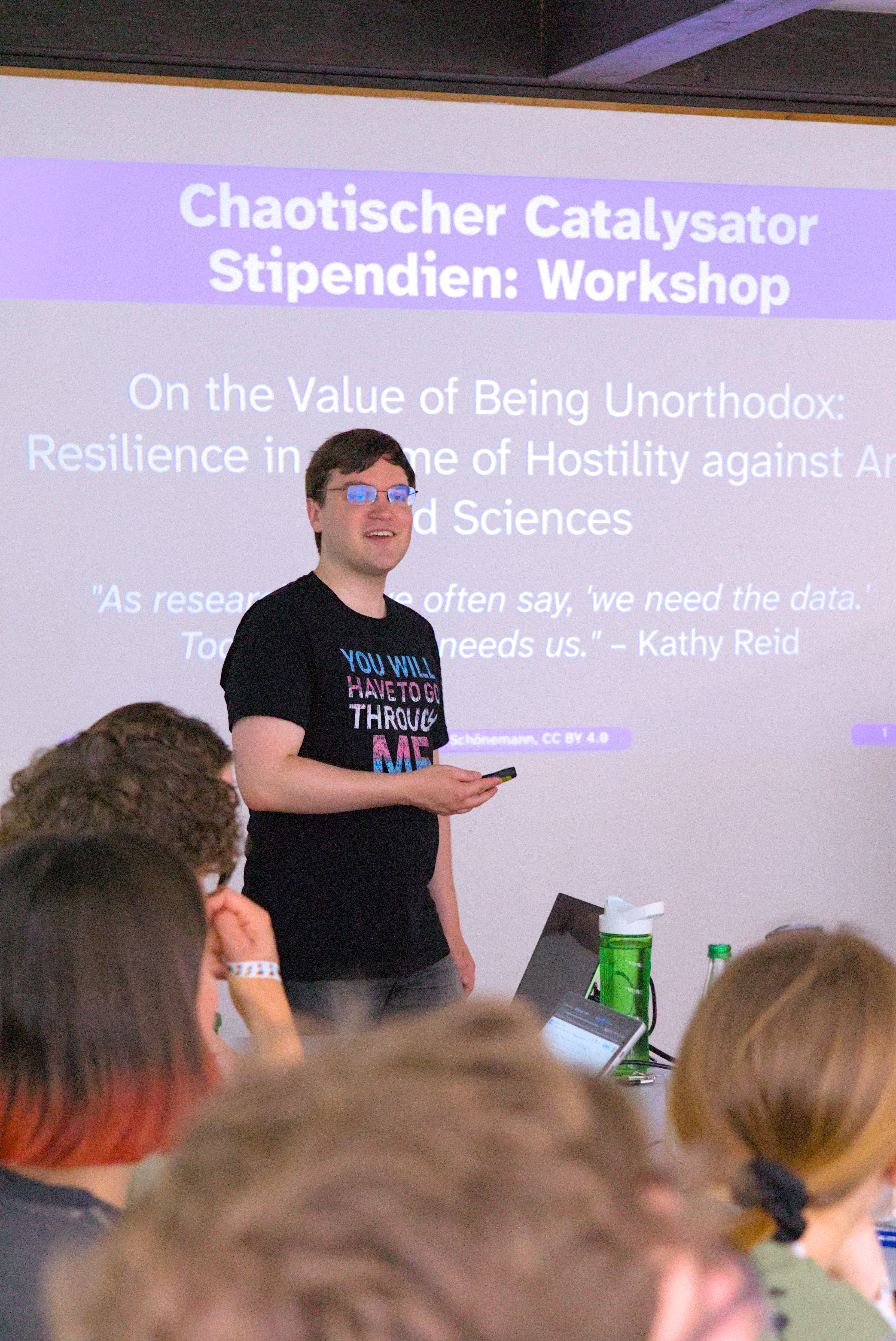 Henrik steht mit einer Fernbedinung in der Hand neben einem Projektor. Die Folie im Hintergund zeigt die Begrüßung mit der Folie: Chaotischer Catalysator Stipendium Workshop. Nah an der Kamera sind die Köpfer der Teilnehmenden verschwommen zu erkennen.