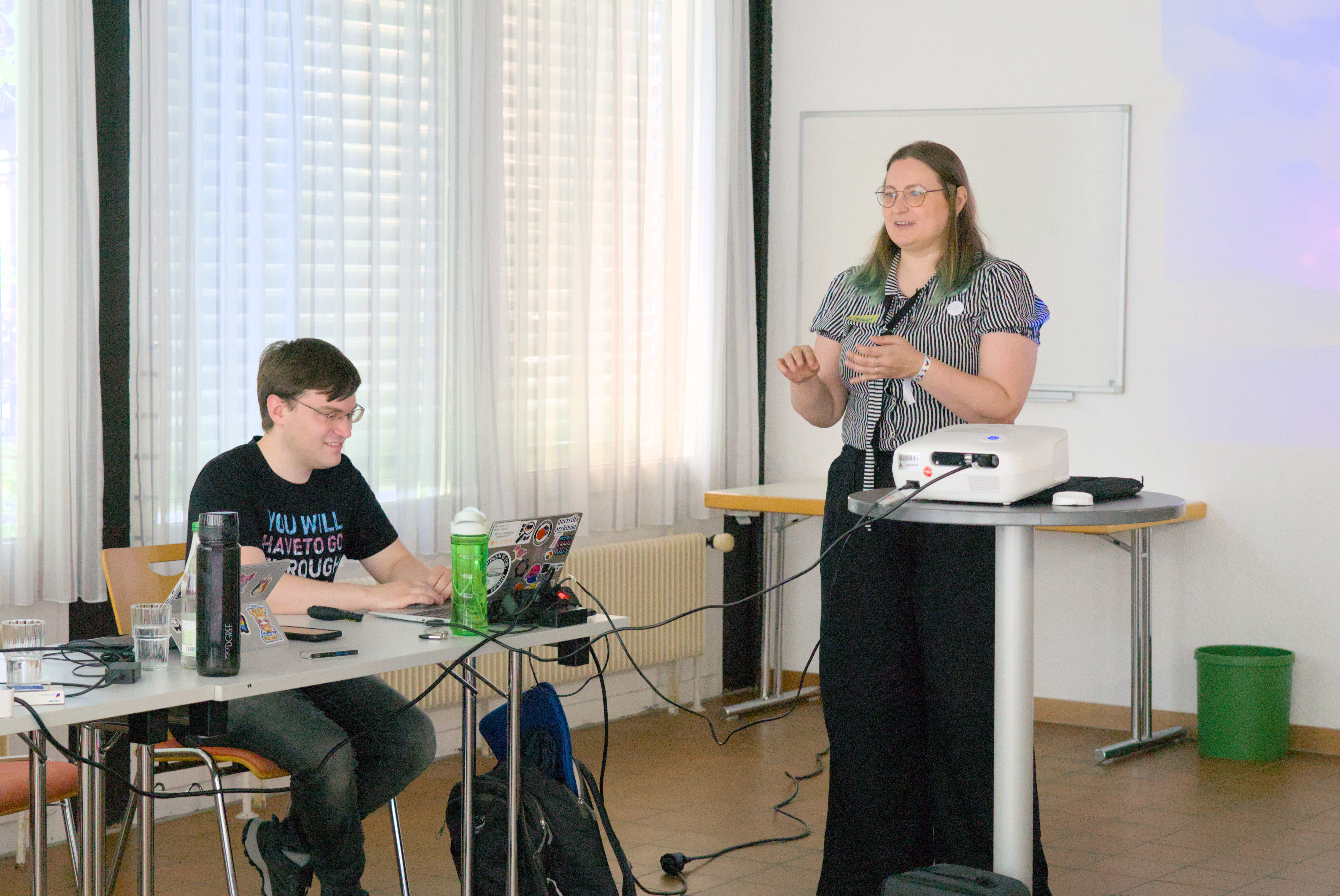 Claudia Präsentiert stehend nicht erkenntliche Folien. Henrik sitzt daneben an seinem Laptop.