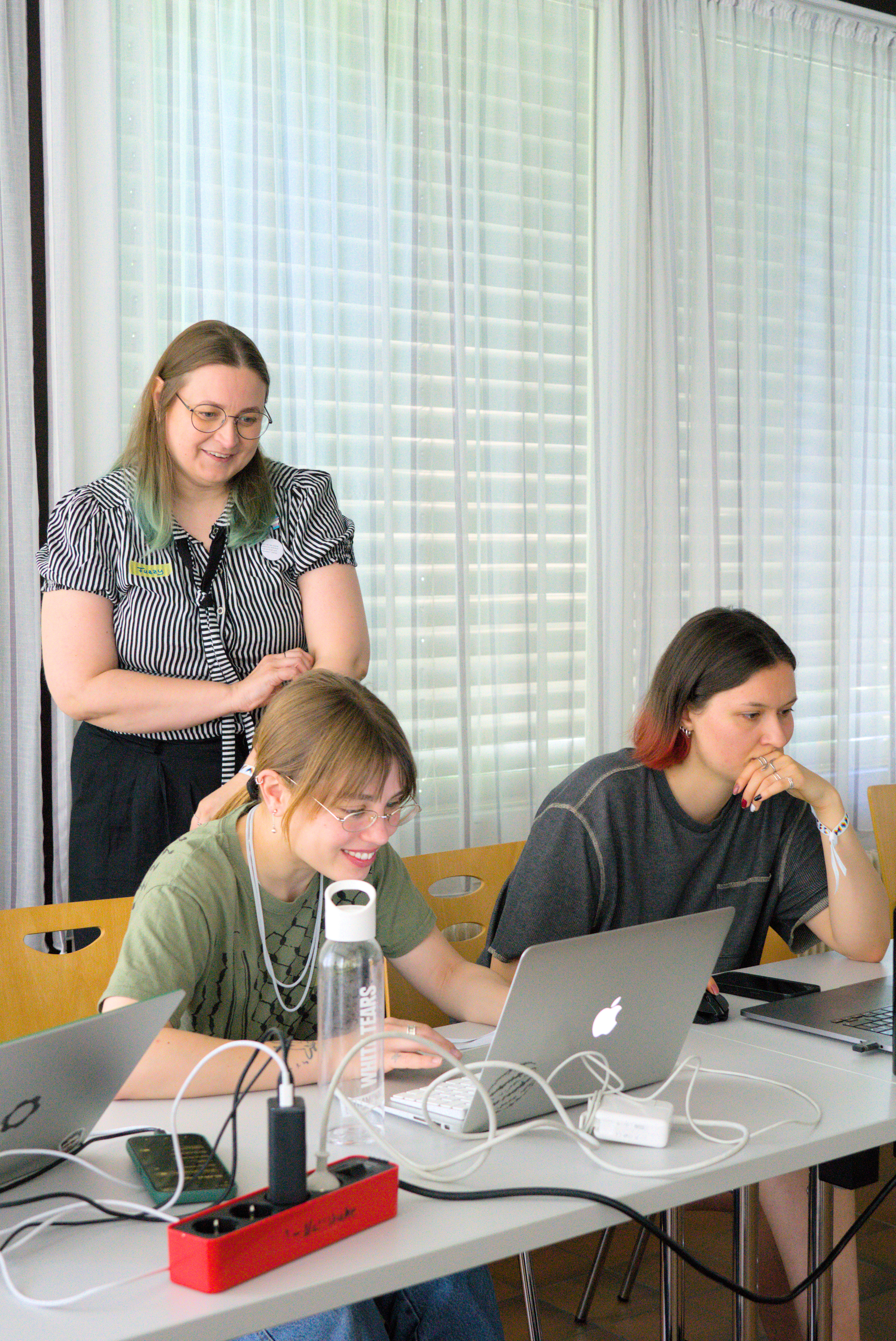 Claudia schaut zwei Teilnehmerinnen von hinten über die Schulter. Die Teilnehmerinnen schauen auf ihren Laptop und sprechen mit Claudia.