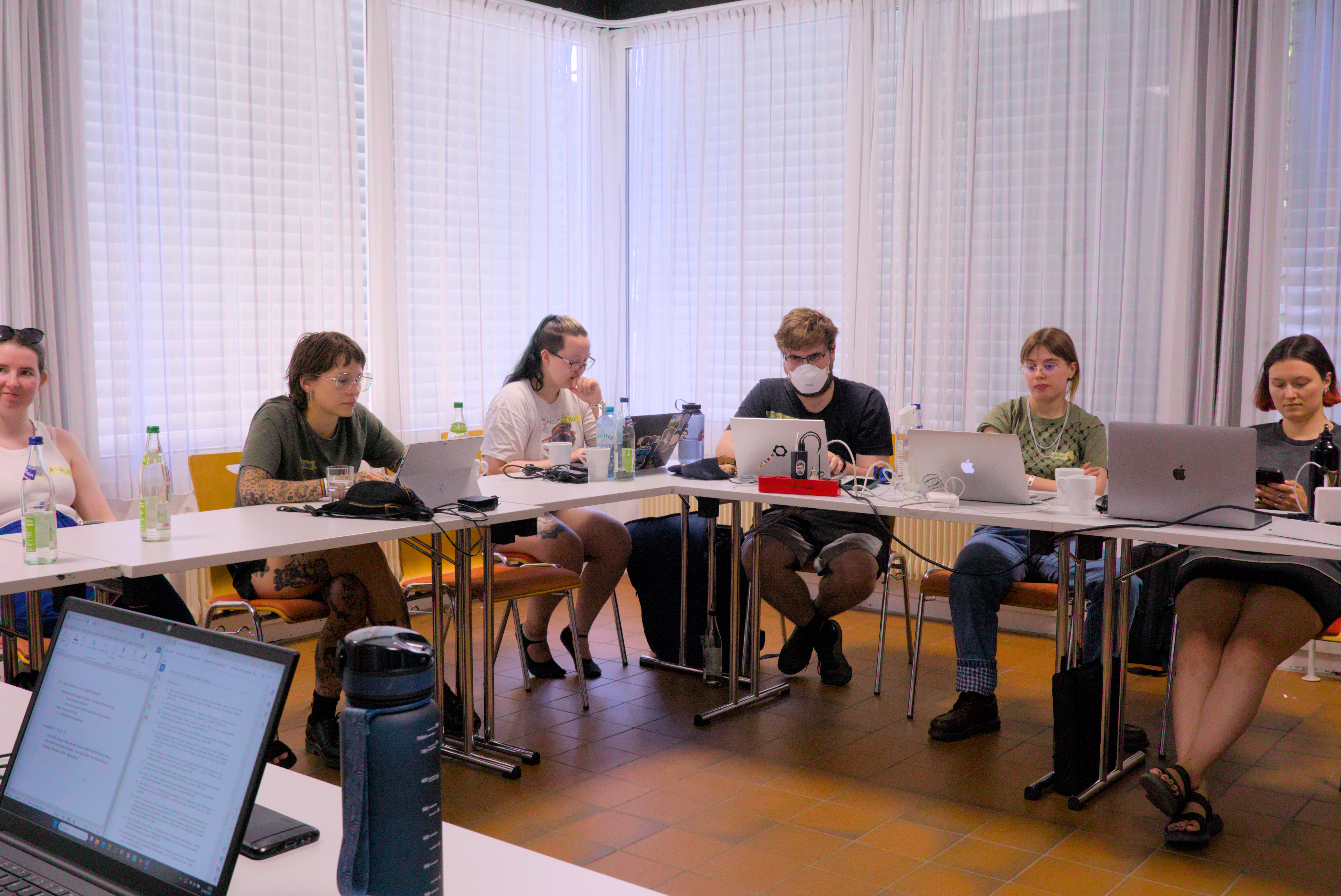 Eine Gruppe Teilnehmer*innen sitzt in U-Form an Tischen. Sie schauen auf ihre Laptops.