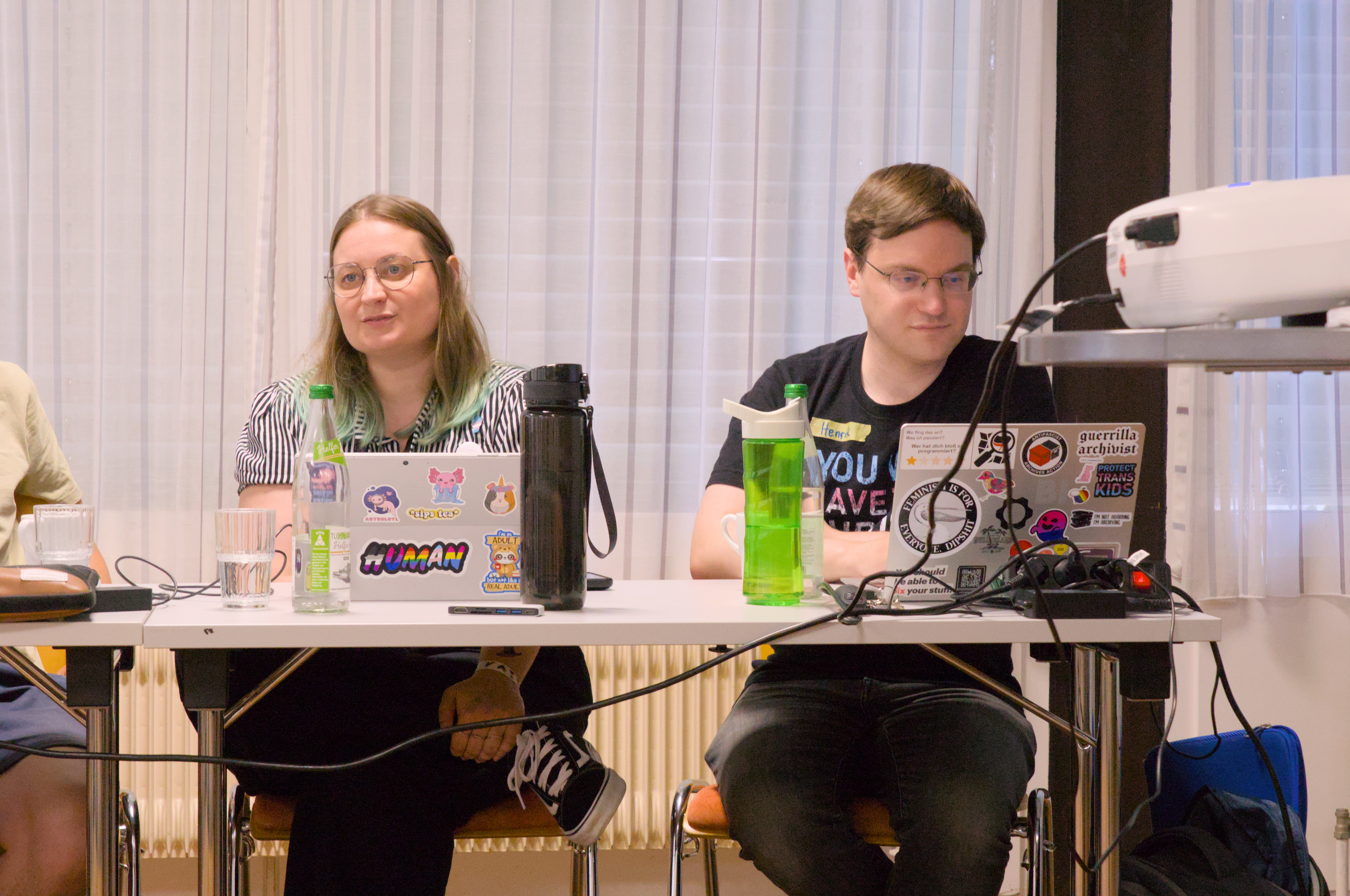Claudia und Henrik sitzen an einem Tisch. Vor ihnen haben sie ihre mit bunten Stickern beklebte Laptops uns Wasserflaschen stehen.